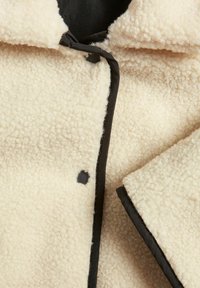 Krämfärgad fleecejacka med en texturerad yta, svarta detaljer och en framknäppning med en toggel. Accenter inkluderar en synlig svart knapp.