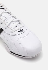 Λευκά δερμάτινα αθλητικά παπούτσια με στρογγυλή μύτη, μαύρη ρίγα και λευκές κορδόνες. Διαθέτει ραμμένο σχέδιο και μαύρο λογότυπο στη γλώσσα.