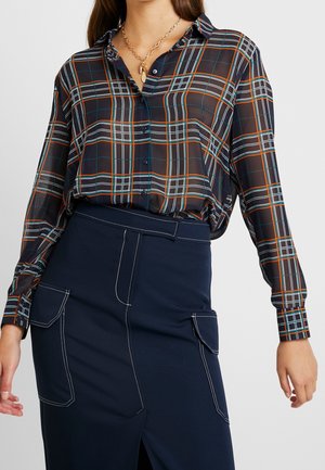 Vrouw draagt een doorschijnend, geruit overhemd met lange mouwen, ingestopt in een marineblauwe rok met witgestikte zakken en een voorsplit.