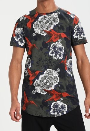 Ciemny t-shirt z krótkim rękawem, z czerwonym kolibrem i białymi kwiatowymi wzorami, noszony przez stojącą osobę z rozluźnionymi ramionami.
