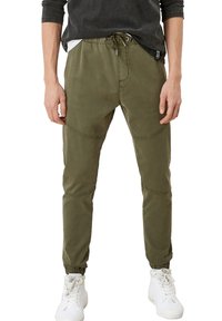 Man in olijfgroene joggerbroeken met elastische enkels en witte hoge sneakers, staande tegen een effen witte achtergrond.