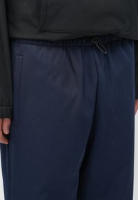 Ναυτικό μπλε ελαφριά παντελόνια με ελαστική μέση, που διαθέτουν πλαϊνή τσέπη και απαλή υφή. Μερικώς καλυμμένα από ένα μαύρο μπουφάν.