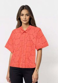 Korallfarget kortermet skjorte med øyeletdesign, knappestolpe og krage. Brukt med svarte bukser, noe som skaper en fargekontrast.