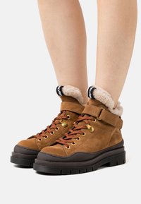 Stivaletti alla caviglia in suede marrone con una morbida fodera color crema, lacci rossi e accessori neri. Presentano una suola in gomma spessa e una cinghia regolabile.