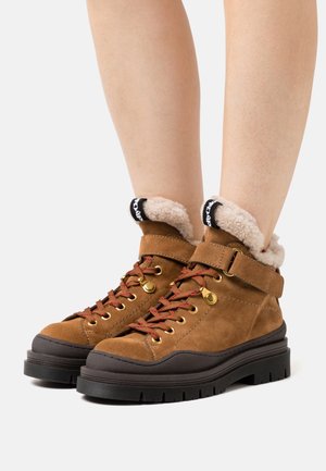 Brązowe zamszowe botki za kostkę z puszystym kremowym wnętrzem, czerwonymi sznurówkami i czarnymi detalami. Posiadają grubą gumową podeszwę i regulowany pasek.