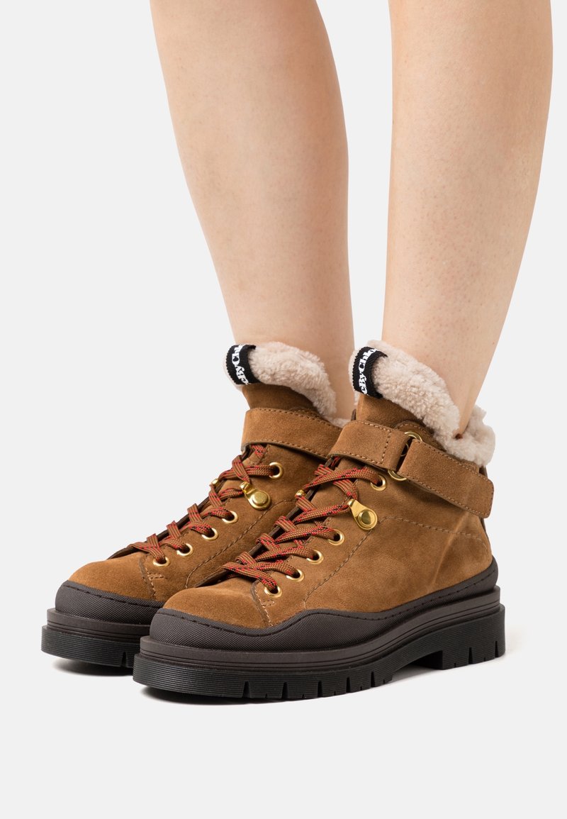 Stivaletti alla caviglia in suede marrone con una morbida fodera color crema, lacci rossi e accessori neri. Presentano una suola in gomma spessa e una cinghia regolabile.