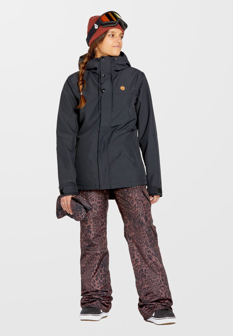 Schwarze wasserdichte Jacke mit Reißverschluss, kombiniert mit braunen Leopardmuster-Schneehosen, und accessoriert mit einer kastanienbraunen Mütze und einer Schutzbrille.