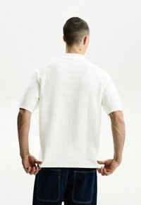 Weißes, strukturiertes Poloshirt mit kurzen Ärmeln und einem weichen Kragen. Die Person ist von hinten zu sehen und passt den Saum an, während sie blaue Jeans trägt.