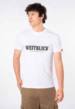Young man with curly hair wearing a white T-shirt with mountains and "WEITBLICK" text, paired with brown cargo shorts.