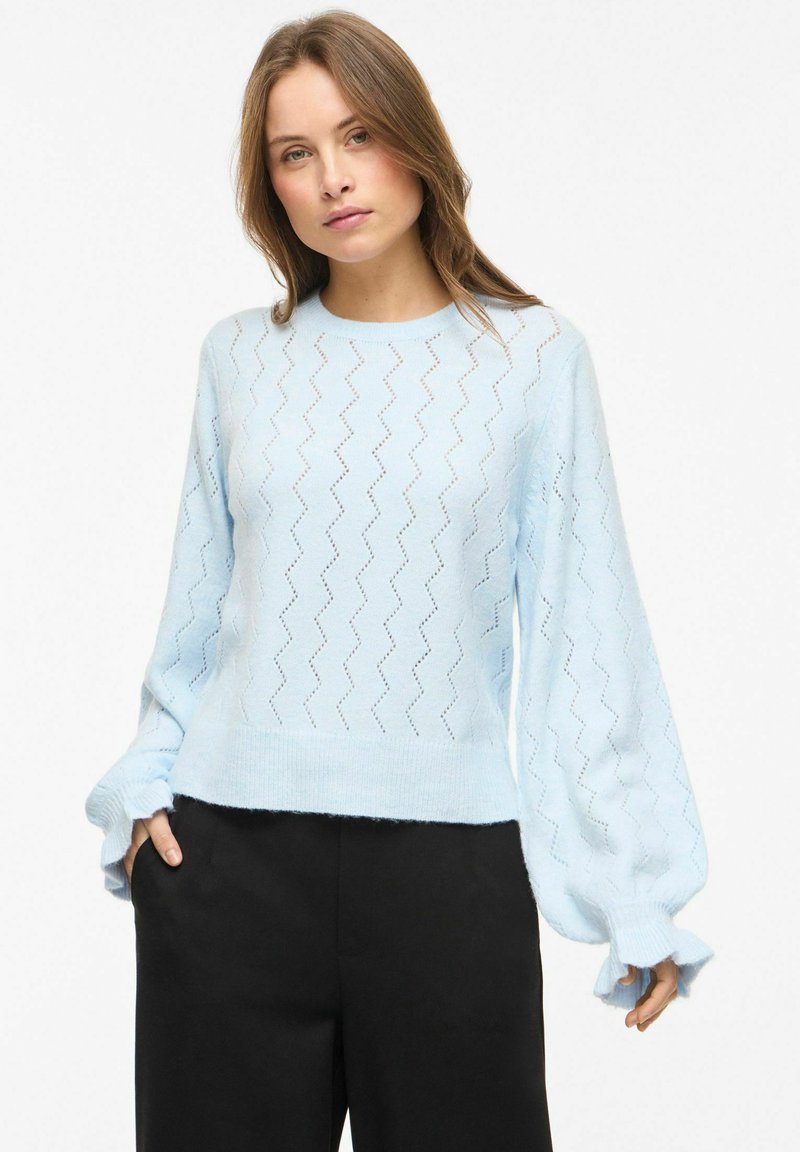 Femme portant un pull en tricot bleu clair avec un motif en zigzag et des manches bouffantes, assorti à un pantalon noir, debout devant un fond uni.