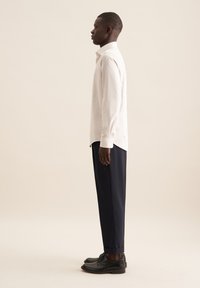 Camicia a righe bianche e rosa chiaro, slim fit. Pantaloni blu scuro, taglio sartoriale. Scarpe nere, profilo basso, texture liscia. Vista di profilo in piedi.