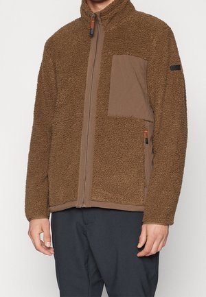Brązowa polarowa kurtka na zamek błyskawiczny z fakturowanej tkaniny, z pikowaną kieszenią na piersi, boczną kieszenią na zamek, noszona przez stojącą osobę w ciemnych spodniach.