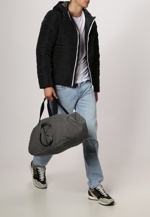 Bolso de viaje tejido en gris con forma redondeada, acentos negros y dos asas resistentes; lo lleva una persona vestida de manera informal.