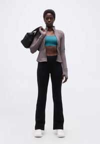 Αθλητική γυναίκα με πετρόλ αθλητικό μπουστάκι, γκρι ζακέτα με φερμουάρ, μαύρο παντελόνι και λευκά αθλητικά παπούτσια, κρατώντας μια μαύρη τσάντα γυμναστηρίου στον ώμο.