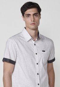Camisa de manga corta abotonada en tejido gris claro con un patrón texturizado, puños de acento azul oscuro y un bolsillo en el pecho con el logo.