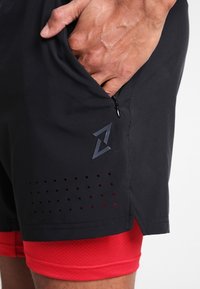 Svarta sportshorts med en dragkedjeficka, med en strukturerad röd foder och perforerade detaljer nära nederkanten. Dragkedjeöglan synlig.
