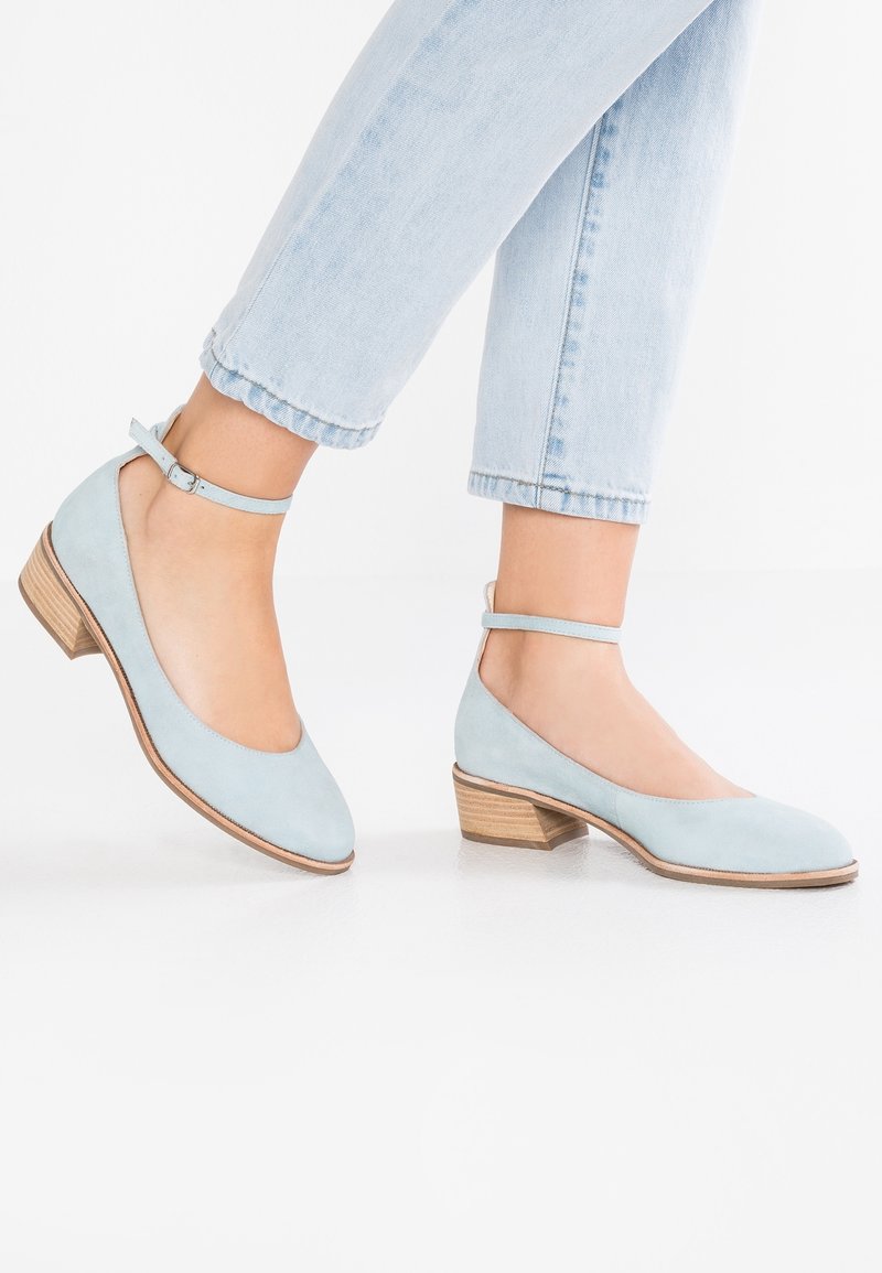 Ballerines en daim bleu clair avec un bout arrondi, une bride à la cheville et un petit talon en bois, portées avec un jean en denim clair retroussé.