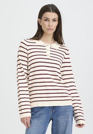 Nő, aki világos krémszínű, hosszú ujjú pulóvert visel sötét vízszintes csíkokkal és gallérral, kék farmerrel, egyszínű háttér előtt.