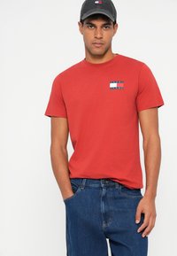 Červené bavlněné tričko s malou námořnickou a bílou nášivkou. V kombinaci s modrými džíny, které mají uvolněný střih a klasický design.