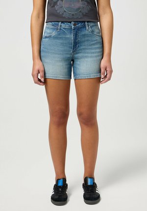 Pantalones cortos de mezclilla en azul claro con un lavado descolorido. Diseño clásico de cinco bolsillos y dobladillos cosidos. Llevados con zapatillas negras con acentos azules.