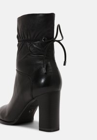 Botte de cheville en cuir noir avec un talon bloc, détail froncé à l'arrière et nœud noué en haut, vue de derrière sur un fond blanc.