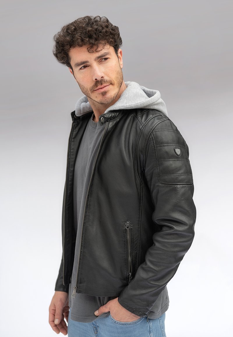 Hombre con cabello rizado que lleva una chaqueta de cuero negra sobre una sudadera con capucha gris y una camiseta, de pie con una mano en el bolsillo.