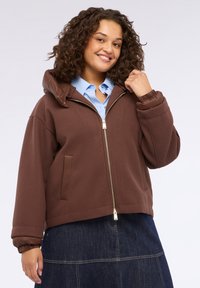 Jachetă cu fermoar maro, cu glugă, având o căptușeală matlasată, manșete elastice și buzunare laterale, purtată peste o cămașă albastră deschisă.