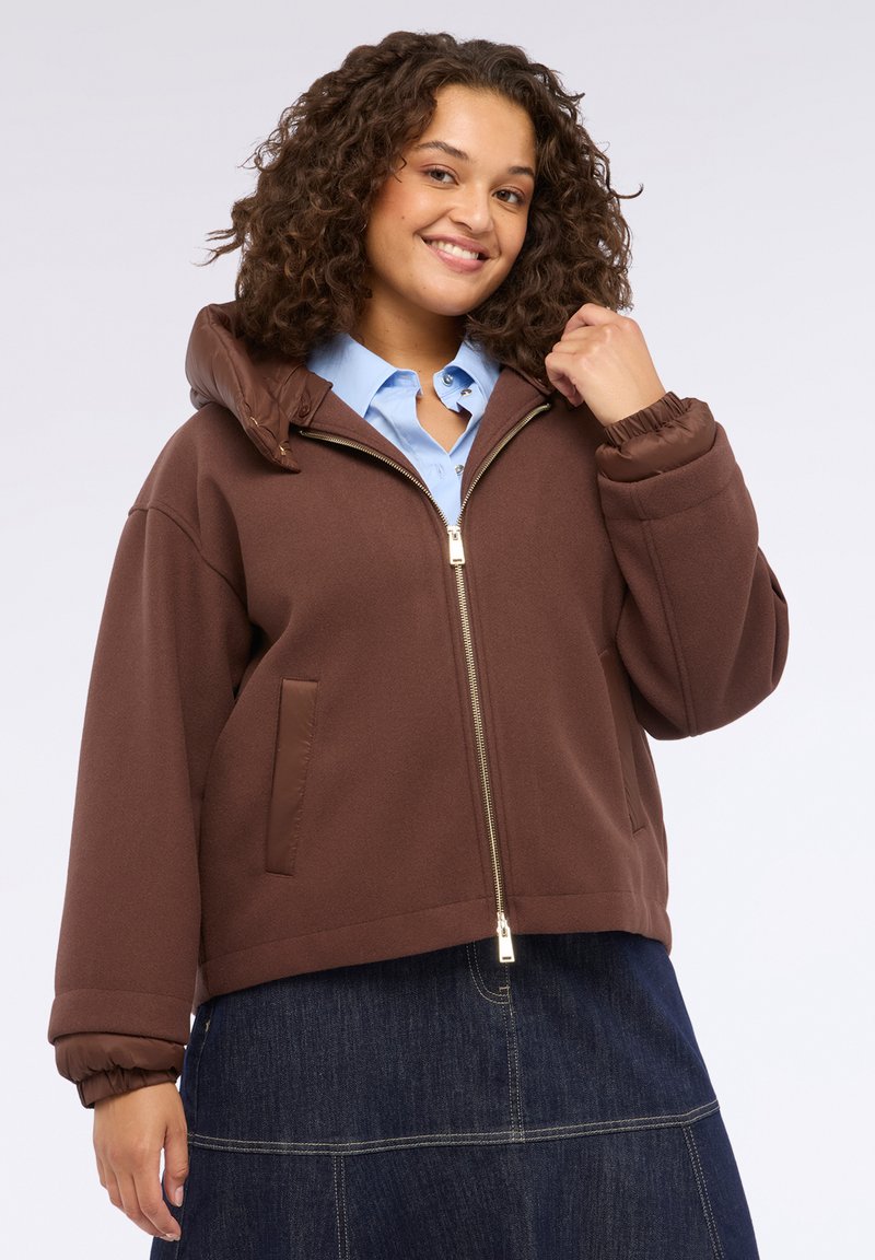 Jachetă cu fermoar maro, cu glugă, având o căptușeală matlasată, manșete elastice și buzunare laterale, purtată peste o cămașă albastră deschisă.