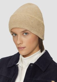 Beanie en tricot côtelé beige fabriqué à partir d'un matériau doux, avec un accent logo brodé. Le design est ajusté et légèrement pointu sur le dessus.