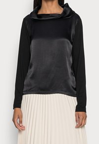 Mustapaita, jossa on pitkät hihat ja satiininen kauluspanta. Materiaali kontrastoi laskostetun, vaalean alaosan kanssa. Suojaava rakenne.