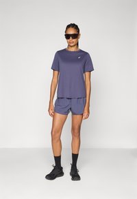 Fialové športové tričko s guľatým výstrihom, v kombinácii s svetlofialovými šortkami. Čierne bežecké topánky a slnečné okuliare dopĺňajú outfit.