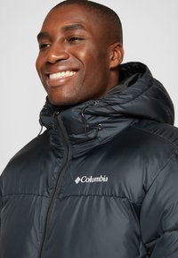 Chaqueta de plumas en azul marino oscuro con cierre de cremallera, capucha ajustable con cordones y logotipo blanco de Columbia en el pecho. Textura suave y brillante.