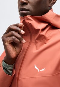 Άτομο με σκουρόχρωμο δέρμα που κλείνει ένα κοραλλί μπουφάν με λευκό γωνιακό λογότυπο στο στήθος.