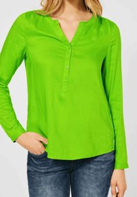 Helle grüne Bluse mit langen Ärmeln, V-Ausschnitt, Knopfleiste, weicher Textur und leichter Raffung an den Schultern. Kombination mit dunklen Jeans.