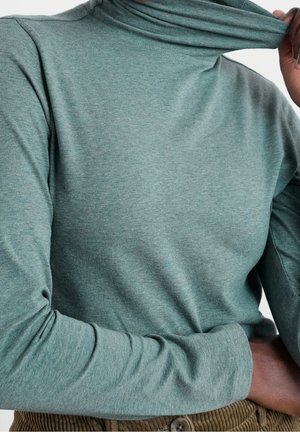 Personne portant un pull vert à col roulé à manches longues, tirant le col vers le haut avec une main, montrant la texture du tissu.