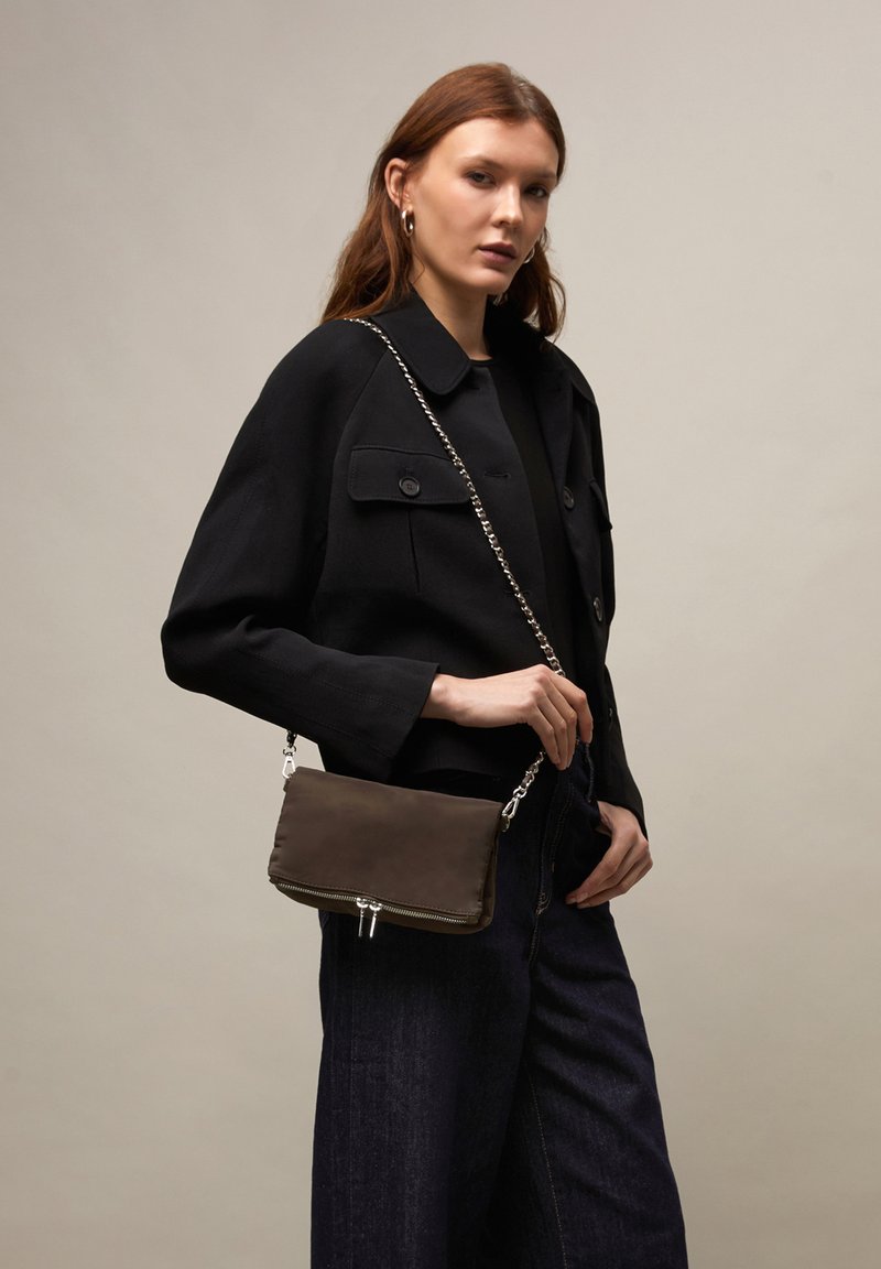 Woman with red hair wears black jacket and dark jeans, holding brown crossbody bag with silver chain strap against a plain beige background.
