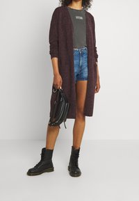 Kvinna som bär en vinröd cardigan, grå crop top, denimshorts och svarta kängor, håller en svart läder crossbody-väska.