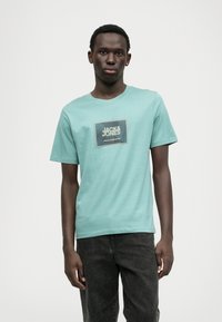 Mätovo zelené bavlnené tričko s obdĺžnikovou grafikou a textom "JACK & JONES" v námorníckej modrej a žltých akcentoch. Okrúhly výstrih, krátke rukávy.