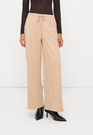 Personne portant un pantalon beige à jambes larges avec une taille à cordon et des bottes noires pointues à talons hauts, debout devant un fond blanc.