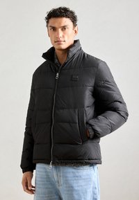 Chaqueta acolchada negra con cuello alto, cierre frontal con cremallera y parche de logo. Presenta puños elásticos y bolsillos laterales, llevada sobre unos jeans de color azul claro.