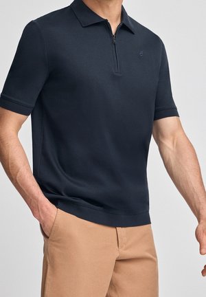 Mann trägt ein dunkelblaues, kurzärmeliges Poloshirt mit Reißverschluss vorne und beige Hose, eine Hand in der Tasche, vor einfachem Hintergrund.