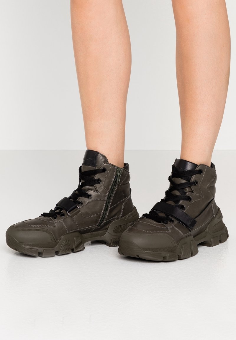 Baskets montantes en vert olive foncé, avec une finition texturée, un système de laçage noir et une fermeture éclair sur le côté pour plus de détails.