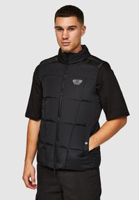 Zwarte pufferweste met hoge kraag, ritssluiting aan de voorkant en geb quilt design. Voorzien van een logopatch en zakken, gedragen over een zwart T-shirt met korte mouwen.