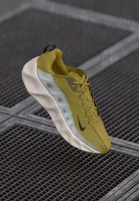 Athletic shoe in olive green with a textured mesh upper, white midsole, and black logo, featuring a rounded toe and lacing system.