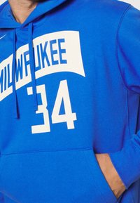 Modrá mikina s kapucí vyrobená z bavlněno-polyesterové směsi, na které je bíle napsáno "MILWAUKEE" a číslo "34". Obsahuje přední kapsu a kapuci se šňůrkou.