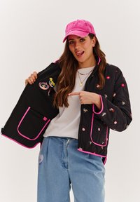 Čierna prešívaná bunda s ružovým lemovaním, so kvetinovými vzormi a nášivkami s kreslenými postavičkami vo vnútri. Nosená s svetlomodrými džínsami a ružovou čiapkou.