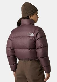 Giacca imbottita viola con colletto alto, caratterizzata da sezioni cucite e un logo bianco sul retro. Un berretto marrone lavorato a maglia completa il look.