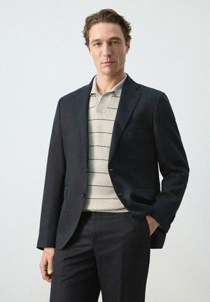 Uomo che indossa un blazer scuro sopra una polo grigia chiara a righe e pantaloni scuri, in piedi con una mano in tasca contro uno sfondo semplice.