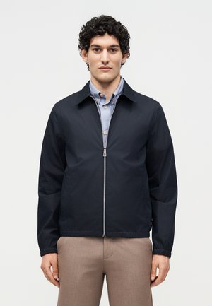 Ung mand med krøllet mørkt hår iført en mørkeblå lynlåstrøje over en lyseblå skjorte med knapper og beige bukser, der står mod en ensfarvet baggrund.