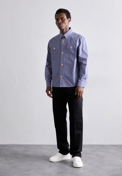 Camisa de botones de manga larga, color azul claro, con dos bolsillos en el pecho y botones dorados, combinada con pantalones negros y zapatillas blancas sobre un suelo texturizado.